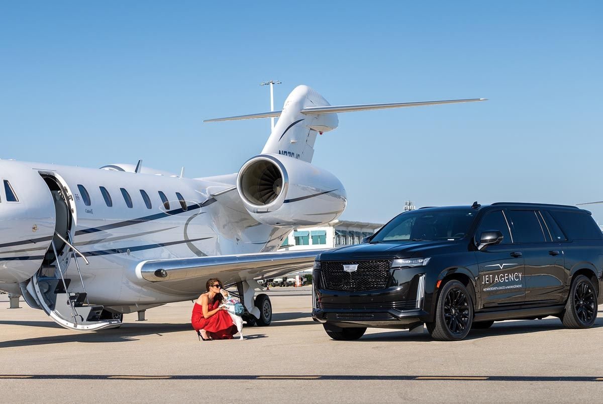A woman in a red dress kneels to hug her dog beside a private jet, a black Jet Agency Escalade parked nearby on the sunlit ramp