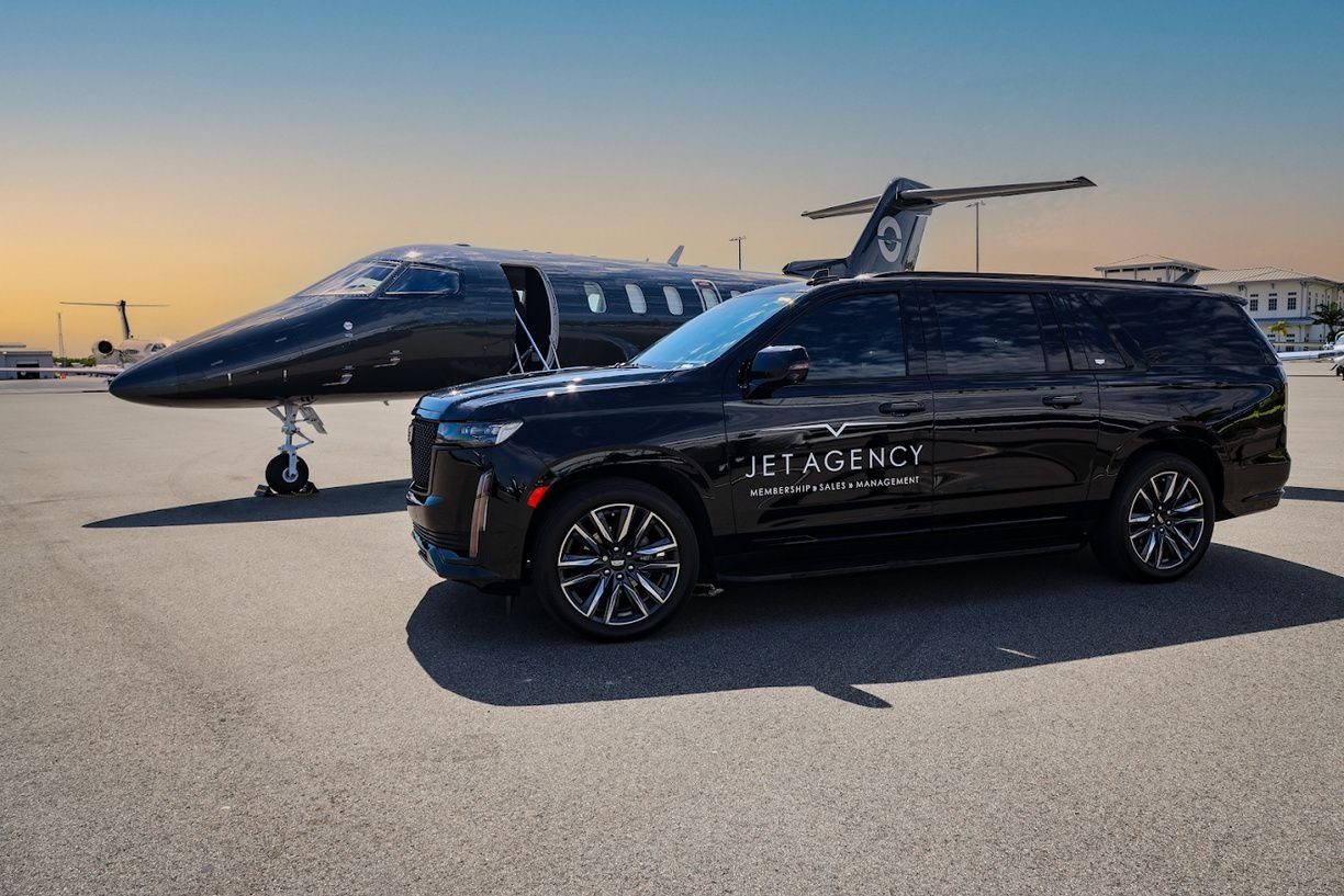 A black Jet Agency Escalade parked beside a black private jet with its door open, on the ramp at sunset